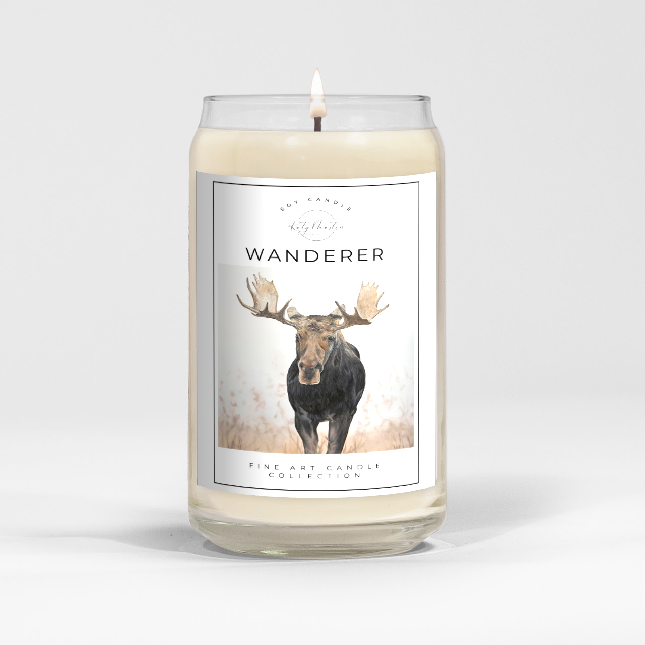 close up of burning white wax candle in a clear glass vessel with fine art landscape painting of a moose by Manitoba Artist Katy Martin on label