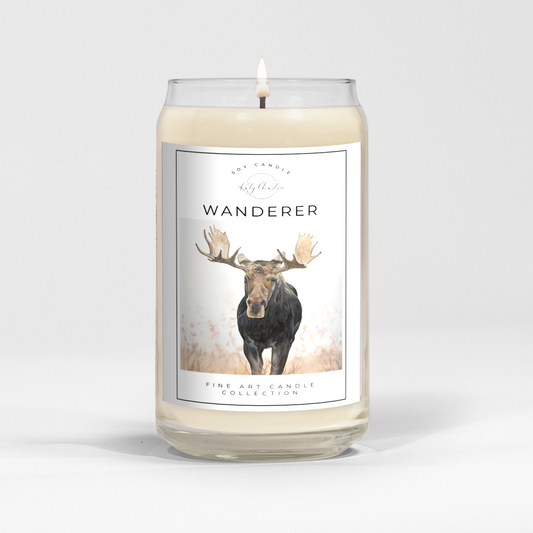close up of burning white wax candle in a clear glass vessel with fine art landscape painting of a moose by Manitoba Artist Katy Martin on label