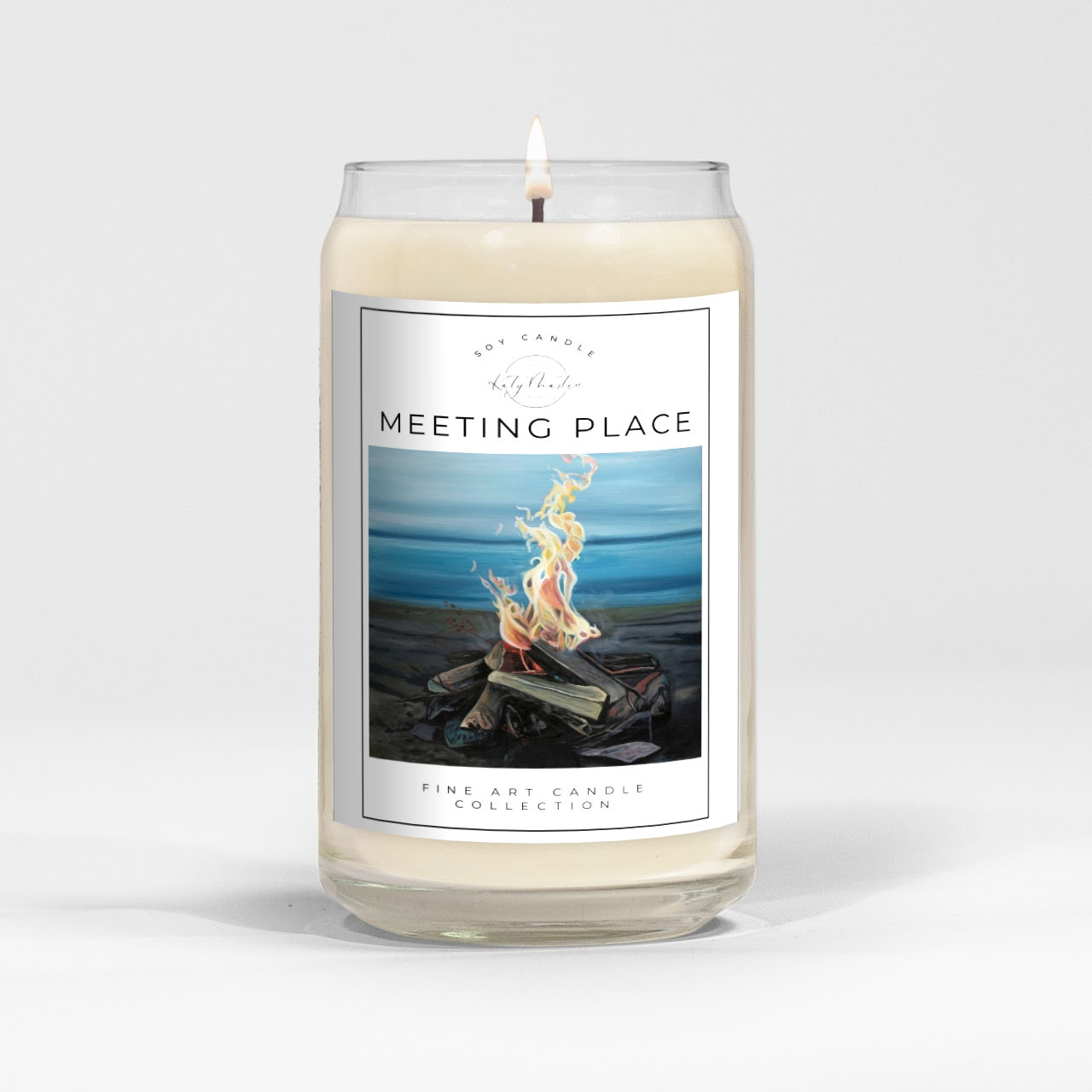 close up of burning white wax candle in a clear glass vessel with fine art landscape painting of a lakeside fire by Manitoba Artist Katy Martin on label