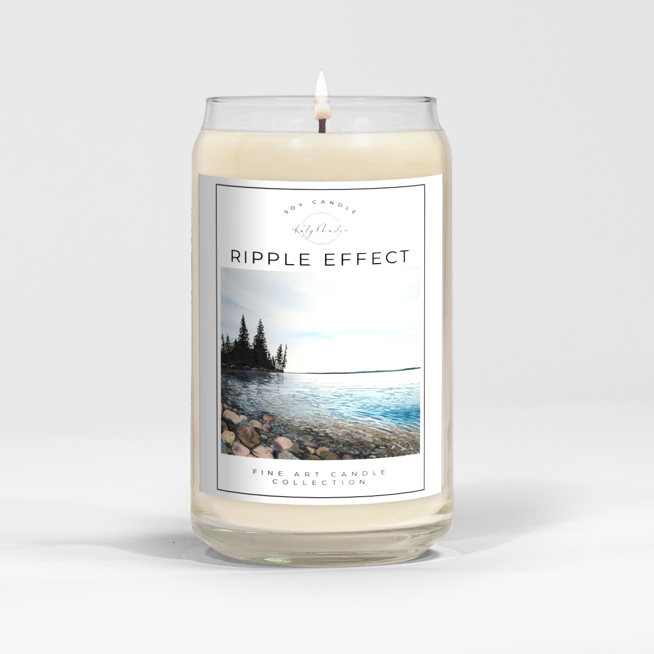 close up of burning white wax candle in a clear glass vessel with fine art landscape painting of clear lake by Manitoba Artist Katy Martin on label