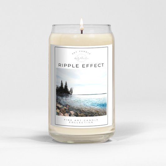 close up of burning white wax candle in a clear glass vessel with fine art landscape painting of clear lake by Manitoba Artist Katy Martin on label