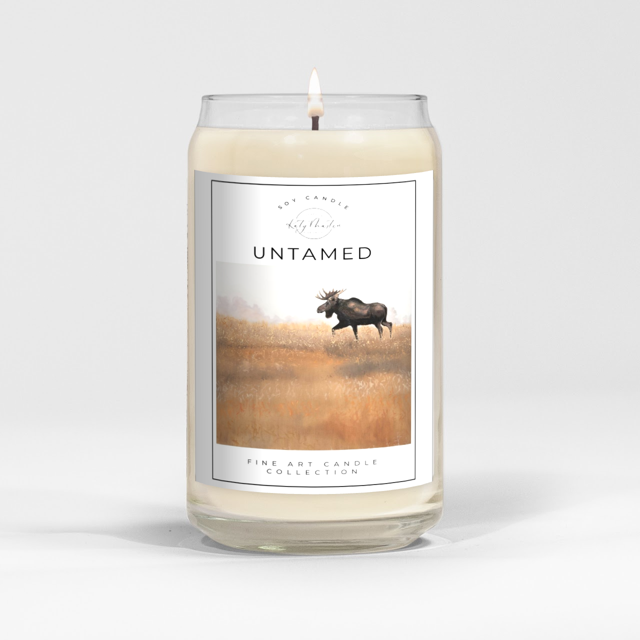 close up of burning white wax candle in a clear glass vessel with fine art landscape painting of a moose by Manitoba Artist Katy Martin on label