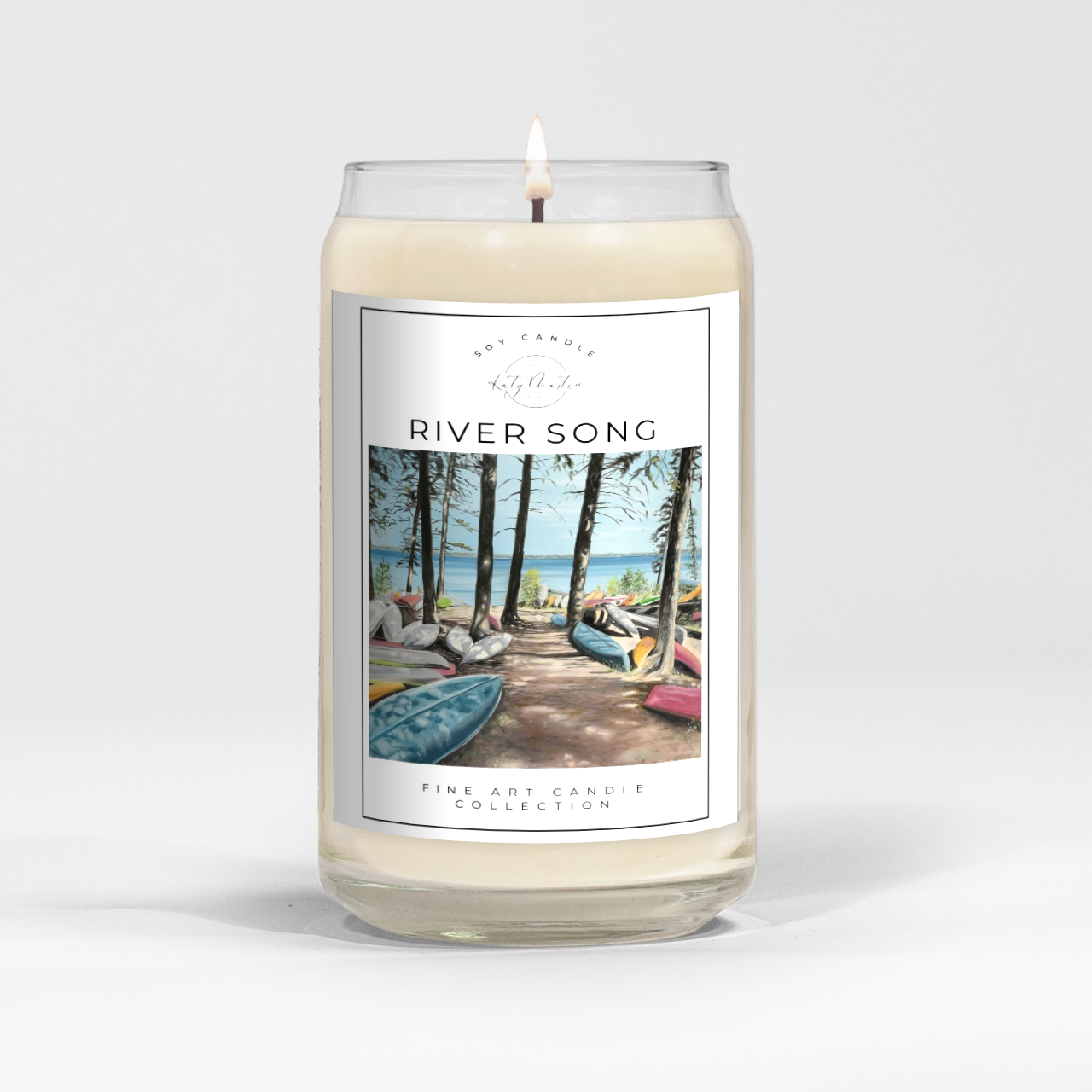 close up of burning white wax candle in a clear glass vessel with fine art landscape painting of canoes at clear lake by Manitoba Artist Katy Martin on label