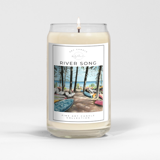 close up of burning white wax candle in a clear glass vessel with fine art landscape painting of canoes at clear lake by Manitoba Artist Katy Martin on label