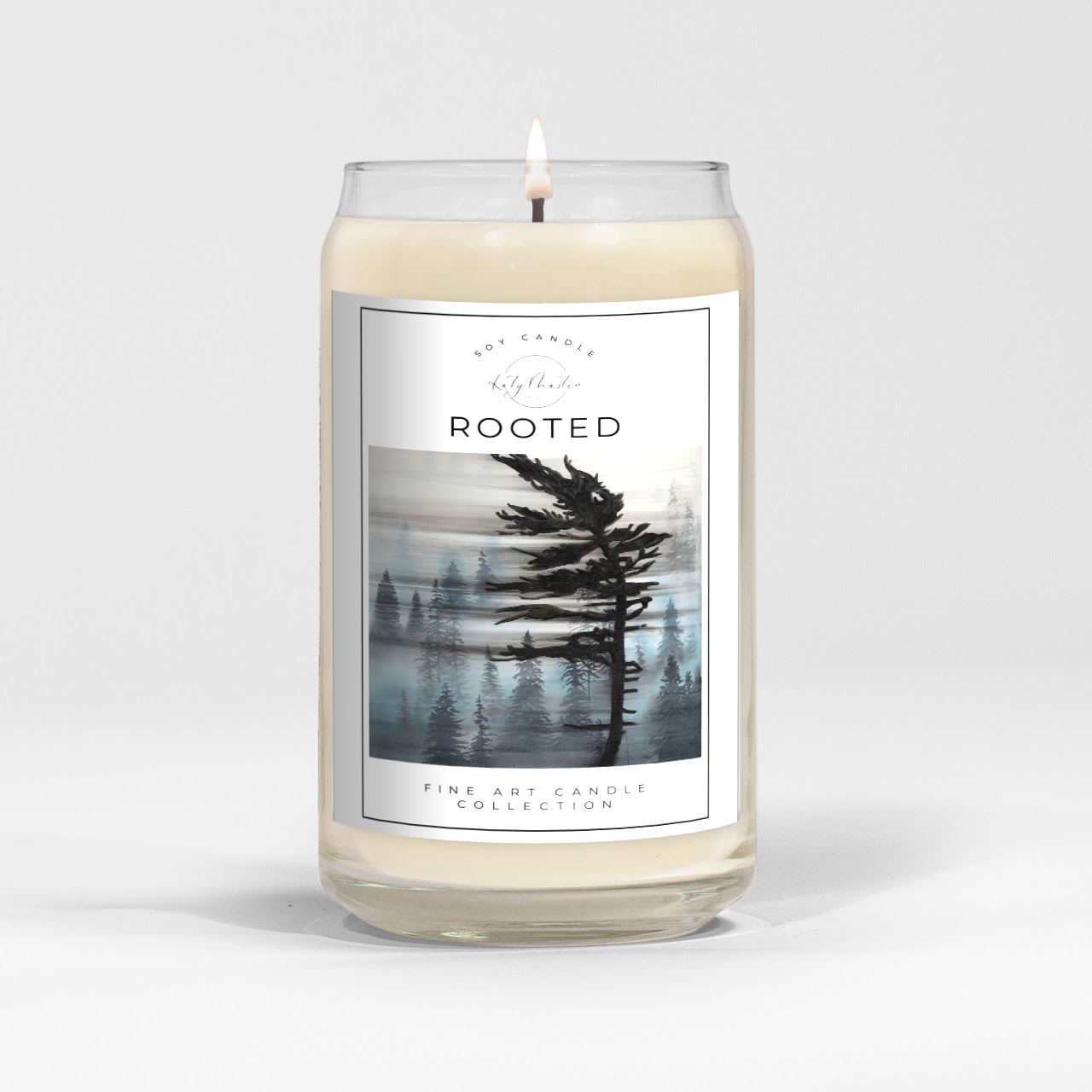 close up of burning white wax candle in a clear glass vessel with fine art landscape painting of a pine tree by Manitoba Artist Katy Martin on label