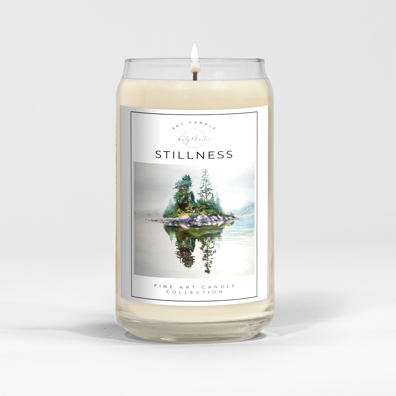 close up of burning white wax candle in a clear glass vessel with fine art landscape painting of an island by Manitoba Artist Katy Martin on label