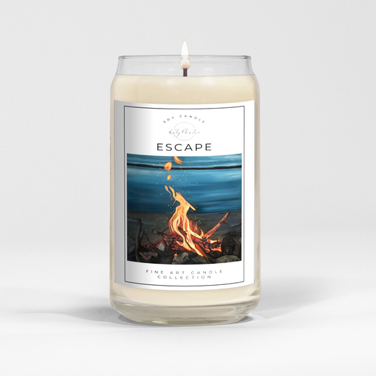 close up of burning white wax candle in a clear glass vessel with fine art landscape painting of a lakeside campfire by Manitoba Artist Katy Martin on label