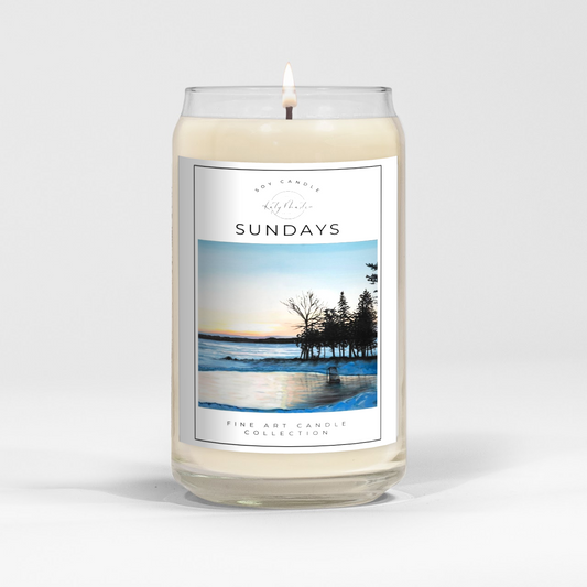 close up of burning white wax candle in a clear glass vessel with fine art landscape painting of an out door hockey rink by a lake by Manitoba Artist Katy Martin on label