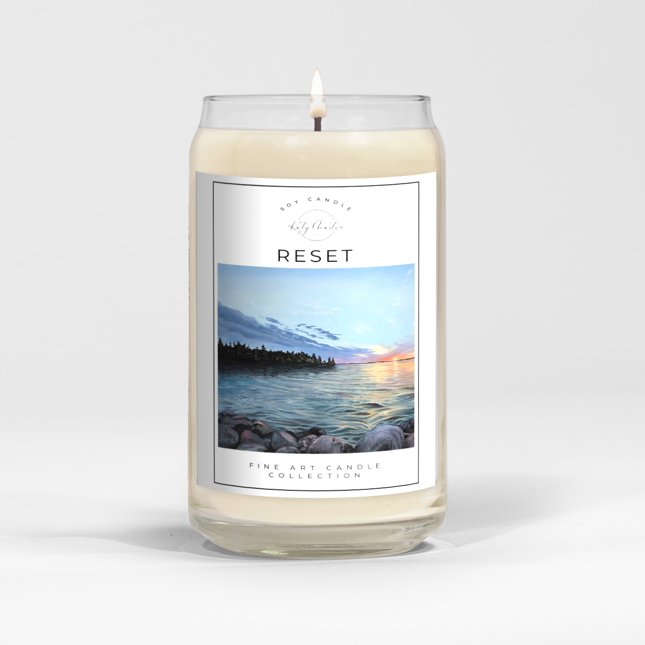 close up of burning white wax candle in a clear glass vessel with fine art landscape painting of a sunset by Manitoba Artist Katy Martin on label