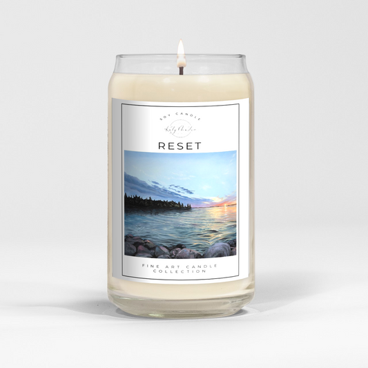 close up of burning white wax candle in a clear glass vessel with fine art landscape painting of a sunset by Manitoba Artist Katy Martin on label