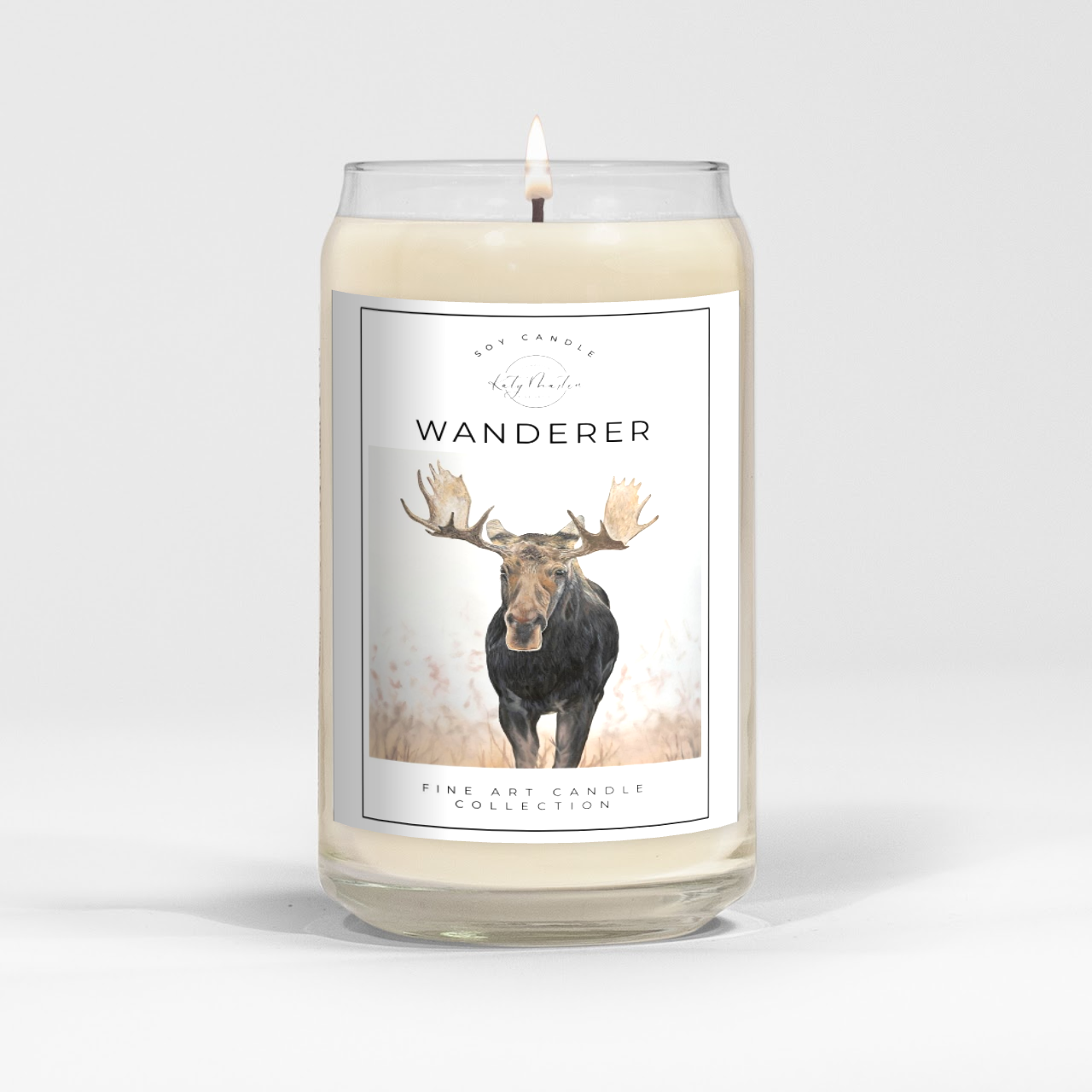 close up of burning white wax candle in a clear glass vessel with fine art landscape painting of a moose by Manitoba Artist Katy Martin on label 