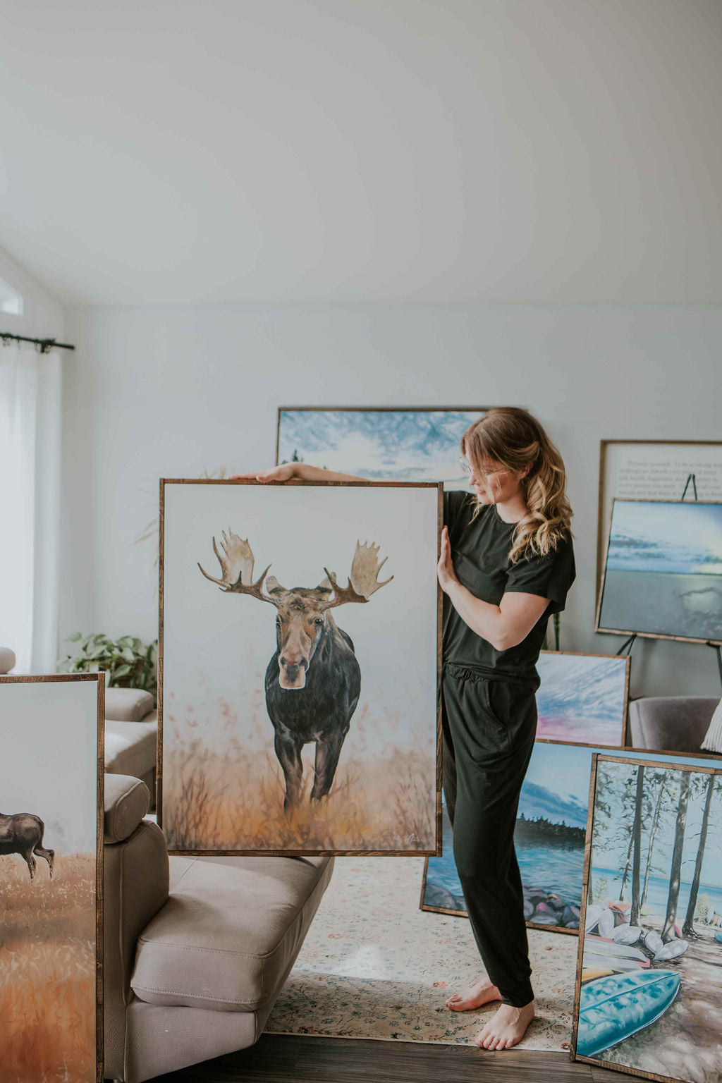 Artist Katy Martin holding her large original painting of a majestic moose titled 'Wanderer' surrounded by her other original paintings from the same collection.